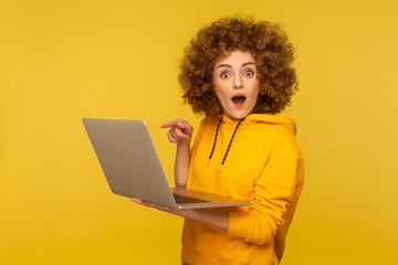 Amazed emotional bright woman with curly hair in urban style hoody pointing laptop screen and looking at camera with shocked expression, open mouth. indoor studio shot isolated on yellow background