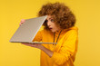 Shocked frightened woman with curly hair in urban style hoody looking furtively at half closed laptop screen, upset scared of error, software failure. indoor studio shot isolated on yellow background
