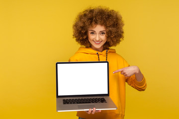 Optimistic happy bright woman with fluffy curly hair in urban style hoody pointing at empty laptop screen, showing mock up, blank display for web ad. indoor studio shot isolated on yellow background