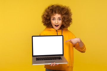 Shocked, surprised woman with fluffy curly hair in urban style hoody pointing at empty laptop screen and looking with amazement, showing mock up, blank display for web ad. indoor studio shot isolated