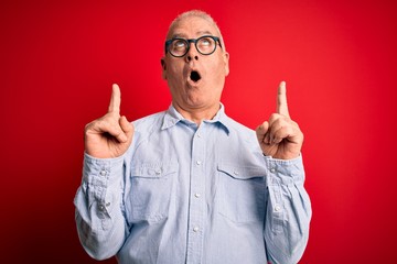 Wall Mural - Middle age handsome hoary man wearing casual striped shirt and glasses over red background amazed and surprised looking up and pointing with fingers and raised arms.