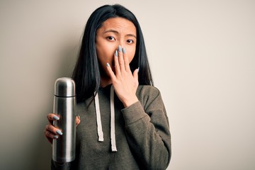 Wall Mural - Young beautiful chinese woman holding thermo standing over isolated white background cover mouth with hand shocked with shame for mistake, expression of fear, scared in silence, secret concept