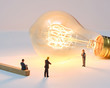 © Sebastian - A businesswoman is lit by a large filament lightbulb (idea) as she presents to colleagues - Tiny People Working From Home