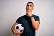 Handsome african american man playing footbal holding soccer ball over white background with hand on chin thinking about question, pensive expression. Smiling with thoughtful face. Doubt concept.
