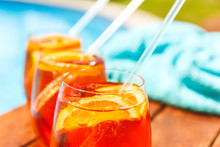 Three Glasses Of Aperol Spritz And Negroni Cold Cocktail With Straws On Wooden Edge Of Swimming Pool With Turquoise Towel. Vacation, Summer, Holiday, Luxury Resort Concept