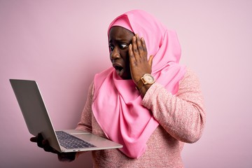 Poster - African american plus size woman wearing muslim hijab using laptop over pink background afraid and shocked, surprise and amazed expression with hands on face