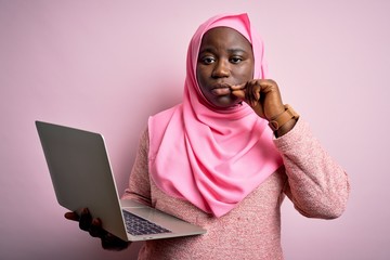 Poster - African american plus size woman wearing muslim hijab using laptop over pink background mouth and lips shut as zip with fingers. Secret and silent, taboo talking