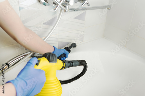cleaning white bathtub and chrome tap with steam generator, hand in blue glove holds yellow steam genetator and hose with brush, which produces steam, home cleaning concept, mold and calcium control