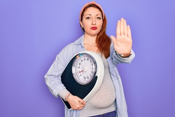 Canvas Print - Young beautiful redhead pregnant woman expecting baby holding weighting machine with open hand doing stop sign with serious and confident expression, defense gesture