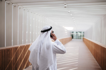 Wall Mural - Emirati guy taking selfie in the urban city lifestyle downtown the heart of Dubai. Arabic guy wearing kandura taking site visit for expo 2020 with investor negotiating over the phone