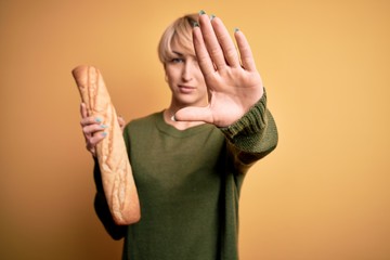 Wall Mural - Young blonde woman with short hair holding fresh bread baguette over yellow background with open hand doing stop sign with serious and confident expression, defense gesture