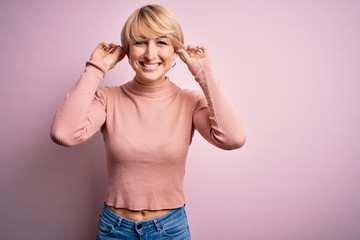 Sticker - Young blonde woman with short hair wearing casual turtleneck sweater over pink background Smiling pulling ears with fingers, funny gesture. Audition problem