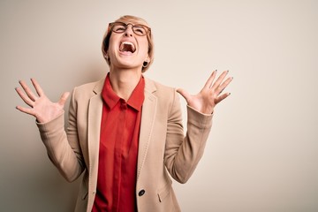 Wall Mural - Young blonde business woman with short hair wearing glasses and elegant jacket crazy and mad shouting and yelling with aggressive expression and arms raised. Frustration concept.