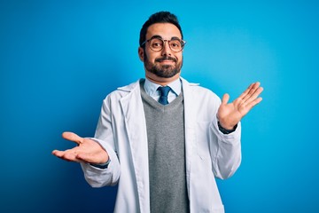 Canvas Print - Young handsome doctor man with beard wearing coat and glasses over blue background clueless and confused expression with arms and hands raised. Doubt concept.