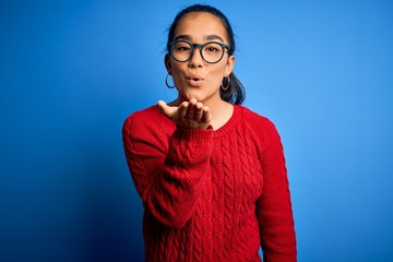 Sticker - Young beautiful asian woman wearing casual sweater and glasses over blue background looking at the camera blowing a kiss with hand on air being lovely and sexy. Love expression.