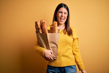 Poster - Young beautiful woman holding a bag of fresh healthy bread over yellow background winking looking at the camera with sexy expression, cheerful and happy face.