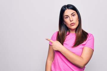 Poster - Young beautiful brunette woman wearing casual pink t-shirt standing over white background Pointing with hand finger to the side showing advertisement, serious and calm face