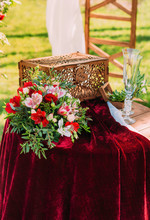 A Beautiful Red Wedding Bouquet Of The Bride Lies On The Table, Champagne Glasses And A Chopped Wooden Box. Outdoor Wedding Ceremony.