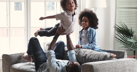 Wall Mural - Happy african ethnic kids siblings doing exercises with strong father on comfortable sofa. Smiling mixed race male parent enjoying gymnastics with teen daughter and preschool son in living room.