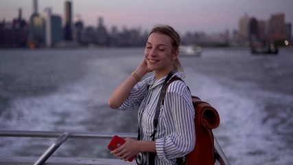 Wall Mural - Slow motion effect of cheerful hipster girl with mobile device for phoning and   camera on neck enjoying boat excursion for exploring New York city, happy Caucasian woman laughing on board
