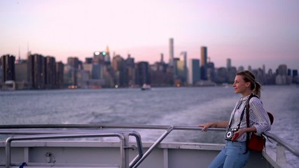 Wall Mural - Smiling hipster traveller with camera technology enjoying evening excursion on water boat for exploring New York megalopolis, happy female tourist with instant camera standing at water boat
