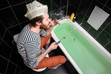 Home Isolation And Quarantine Concept. Holiday At Home, Man Catches Fish In Bath. Newspaper Cap With Unreadable Text