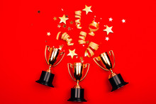 Three Golden Winner Cups With Confetti On A Red Background. Flat Lay Style. Competitions Concept.