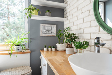 Bathroom with white brick tiles, green plant and window. Wooden counter with ceramic bowl and faucet. Scandinavian interior of bathroom.