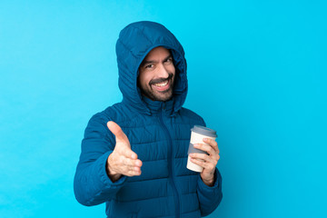 Wall Mural - Man wearing winter jacket and holding a takeaway coffee over isolated blue background shaking hands for closing a good deal