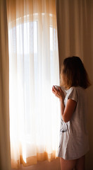 Beautiful young woman in the morning looking at the window and holding a cup of tea or coffee.