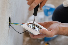 Close Up On Hands Of Caucasian Man Electrician Holding Screwdriver Working On The Plug Electric On Residential Electric System Installing White AC Power Socket On Gray Wall At Home Repair Close Up