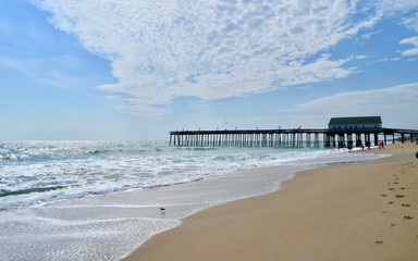  A seascape view of a wooden fishing pier extending into the green foamy waves of the Atlantic Ocean.  Kitty Hawk, Outer Banks, North Carolina.  Copy space.