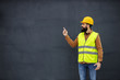 © Dusan Petkovic - Young smiling bearded worker in vest, with helmet on head standing in front of gray wall and pointing up.