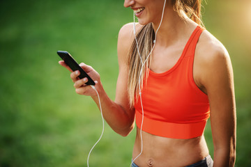 Wall Mural - Cropped picture of young slim charming sportswoman standing in nature, having earphones in ears and using smart phone for choosing music.