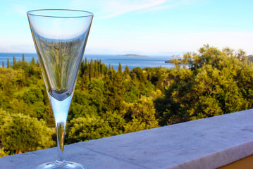 Glass of white wine on seascape background with sun sky mountains and cypress tree landscape reflected in alcohol drink.  An evening cocktail before romantic dinner outdoors on holiday balcony.