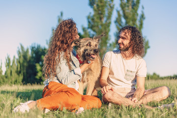 Wall Mural - Happy beautiful curly young couple sits on the grass with with a cute dog in the park