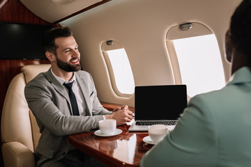 Wall Mural - selective focus of smiling businessman looking at african american businesswoman near laptop with blank screen