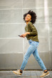 © mimagephotos - Full length side of young black woman walking with mobile phone and earphones