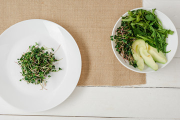 Wall Mural - top view of fresh microgreen on plate near bowl of green salad on beige napkin on white wooden surface