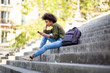 © mimagephotos - Side of young woman sitting on steps with mobile phone and earphones