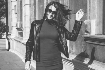 Wall Mural - Brunette woman with a positive smile in a dress and leather black jacket shake hair in street and enjoys a sunny spring day. Lady travel in Batumi, Georgia. Old city at background.