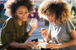 © mimagephotos - two happy young african american women sitting at outdoor cafe with mobile phone and book