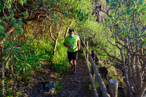 Man with mask running down a path. The corridor is in a forest. The photo was taken in Tenerife, an ideal place to practice trail running. In his hand he has a bottle of water (Soft flask).