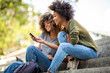 Side of two young african american women sitting on steps outside looking at cellphone