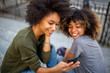© mimagephotos - two happy young black women sitting on steps outside looking at mobile phone