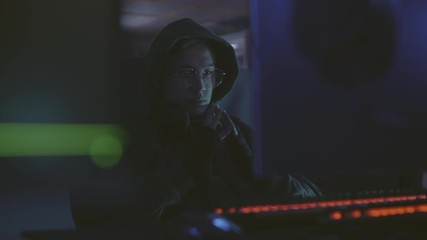 Medium shot of young male program code developer wearing hoody and eyeglasses sitting in front of computer screen and thinking