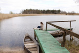 Fototapeta  - Young beautiful woman sits on the wooden bridge on the river at spring day. old boat near the shore. selective focus.