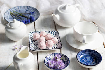 Wall Mural - Rose cream sweet energy balls with coconut flakes in plate, primula and muscaria flowers, tea crockery  on white wooden background