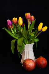 A still life with a bouquet of colourful tulips in a white vase with two pomegranates in front of black background