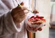 Colazione durante Covid 19. La donna fa colazione tenendo una ciotola con yogurt, cereali e frutti di bosco davanti alla finestra.